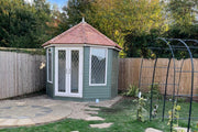 An octagonal-shaped garden room named The Kingsley with a cedar shingle roof, Georgian style doors and windows, situated in a garden setting.