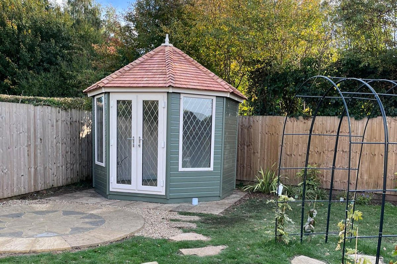 An octagonal-shaped garden room named The Kingsley with a cedar shingle roof, Georgian style doors and windows, situated in a garden setting.