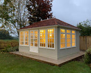 A traditional hip roof garden building with sliding doors and Georgian UPVC windows, illuminated from the inside, set in an outdoor setting.