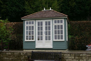 A traditional green garden building with a hip roof, white windows, and a painted door set in a garden environment.