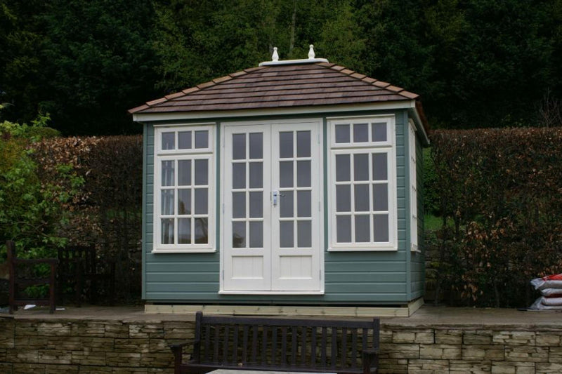 A traditional green garden building with a hip roof, white windows, and a painted door set in a garden environment.